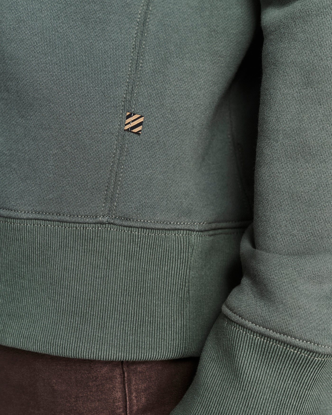 Close-up of a sage green sweatshirt showing the ribbed hem, sleeve cuff, and a small embroidered logo patch on the chest.