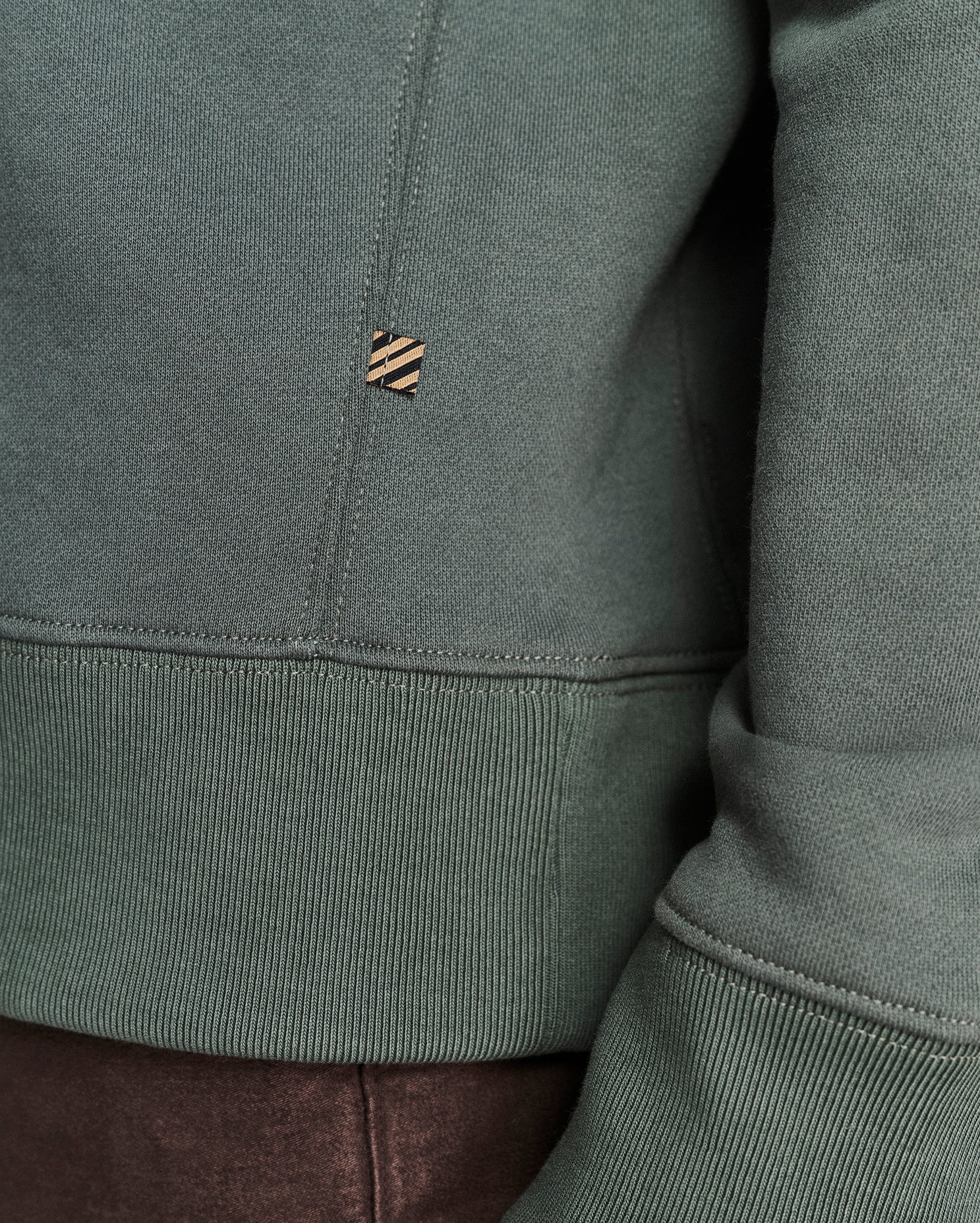 Close-up of a sage green sweatshirt showing the ribbed hem, sleeve cuff, and a small embroidered logo patch on the chest.