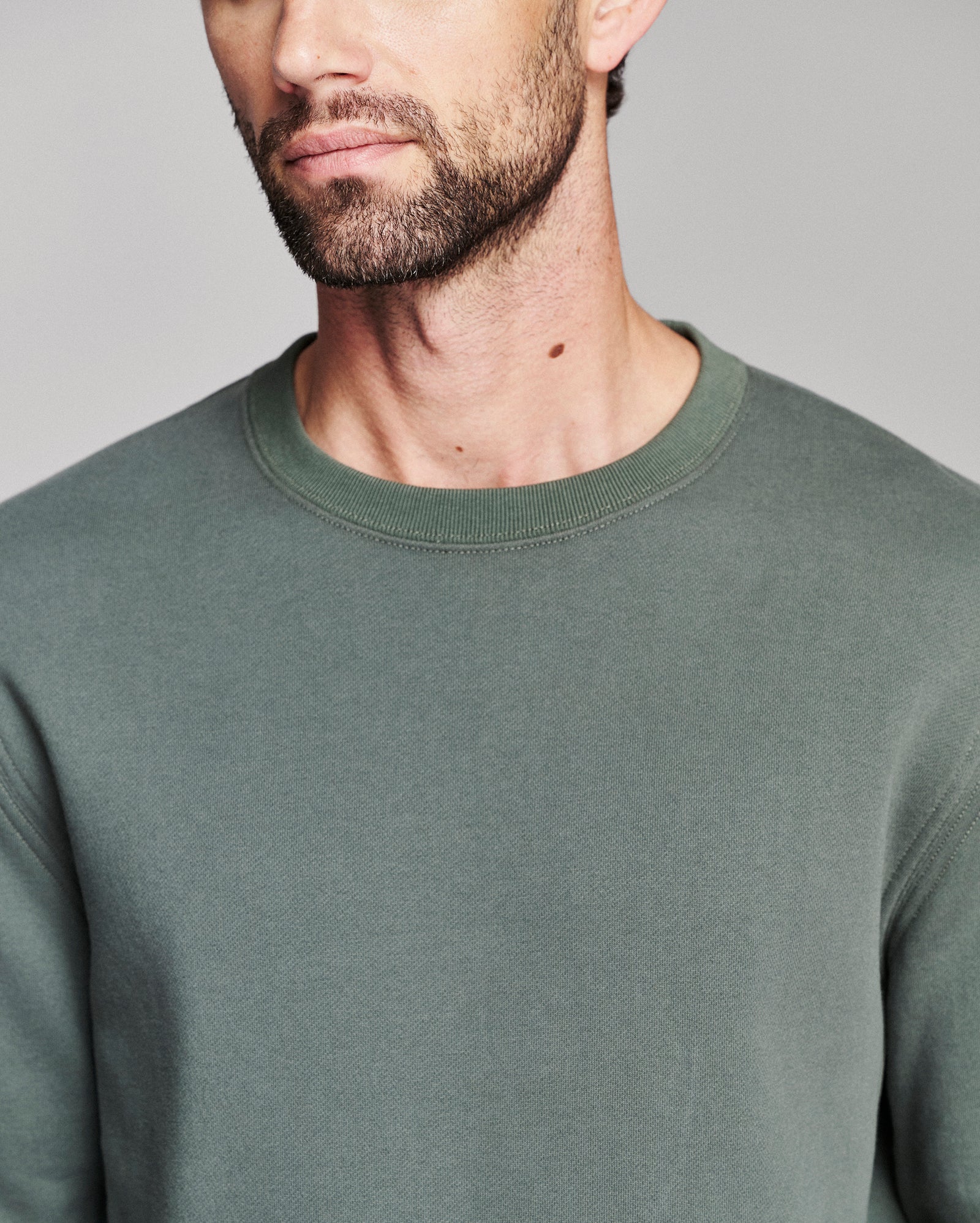 A close-up of a man wearing a muted sage green crewneck sweatshirt against a neutral gray background.