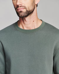 A close-up of a man wearing a muted sage green crewneck sweatshirt against a neutral gray background.