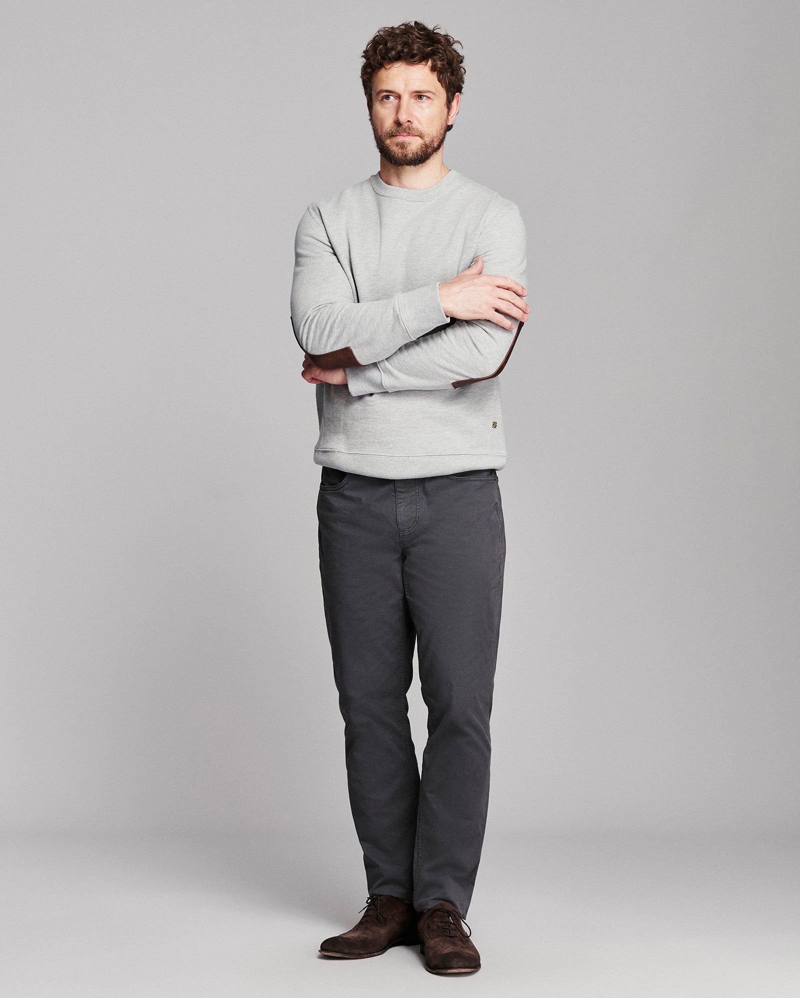 A man wearing a light gray crewneck sweater, dark gray trousers, and brown shoes, standing with arms crossed against a solid gray studio background.