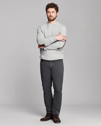 A man wearing a light gray crewneck sweater, dark gray trousers, and brown shoes, standing with arms crossed against a solid gray studio background.