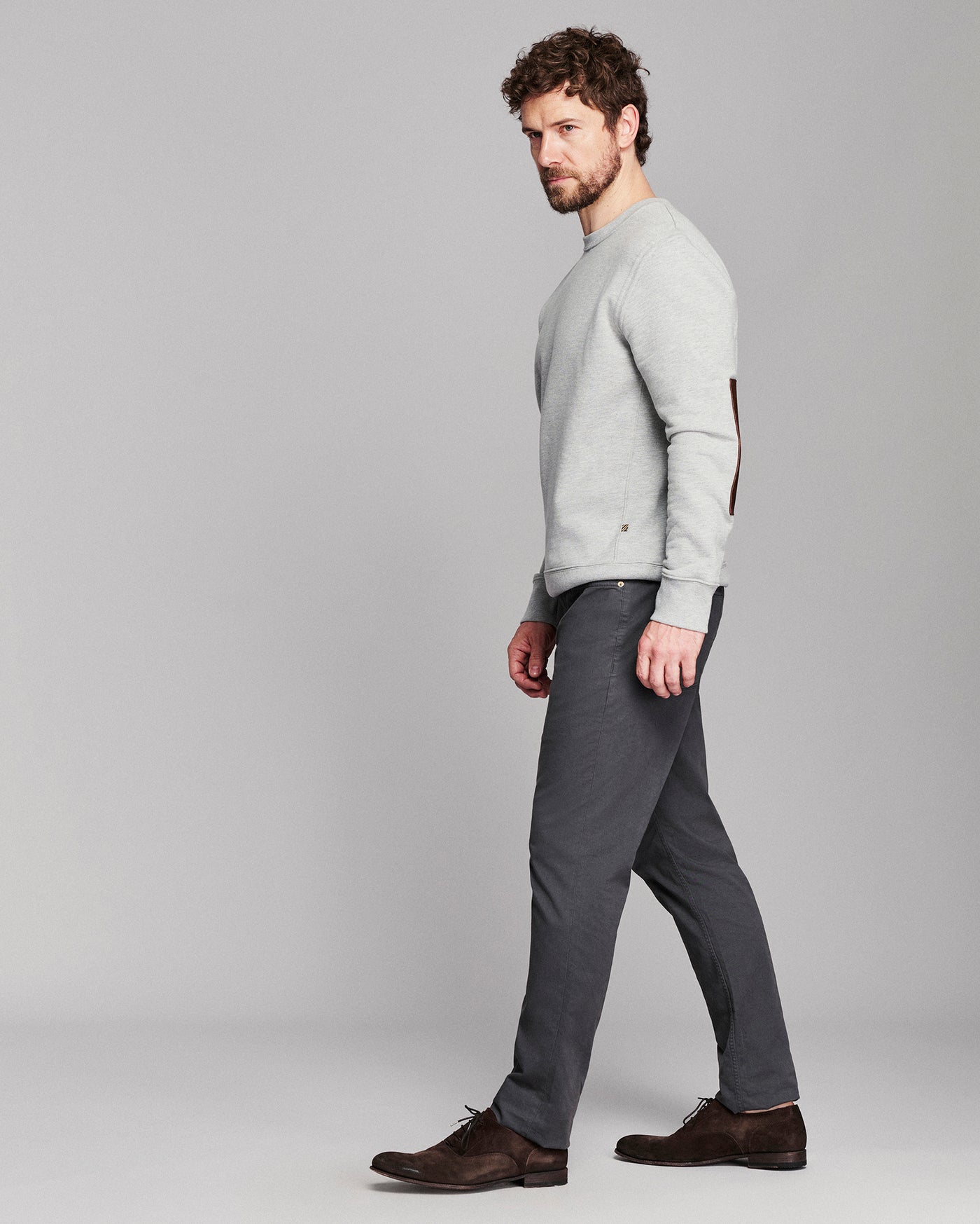 A man in a light gray crewneck sweater, dark gray trousers, and dark brown leather shoes walking in profile against a solid light gray studio background.