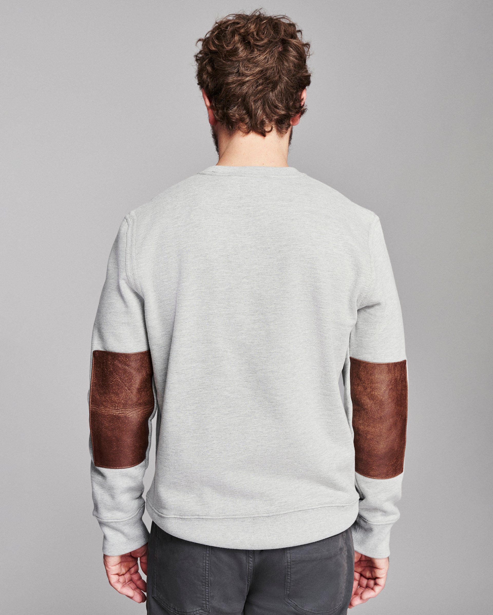 A man viewed from behind wearing a light gray crewneck sweatshirt with brown leather elbow patches, paired with dark gray pants, against a solid gray studio background.