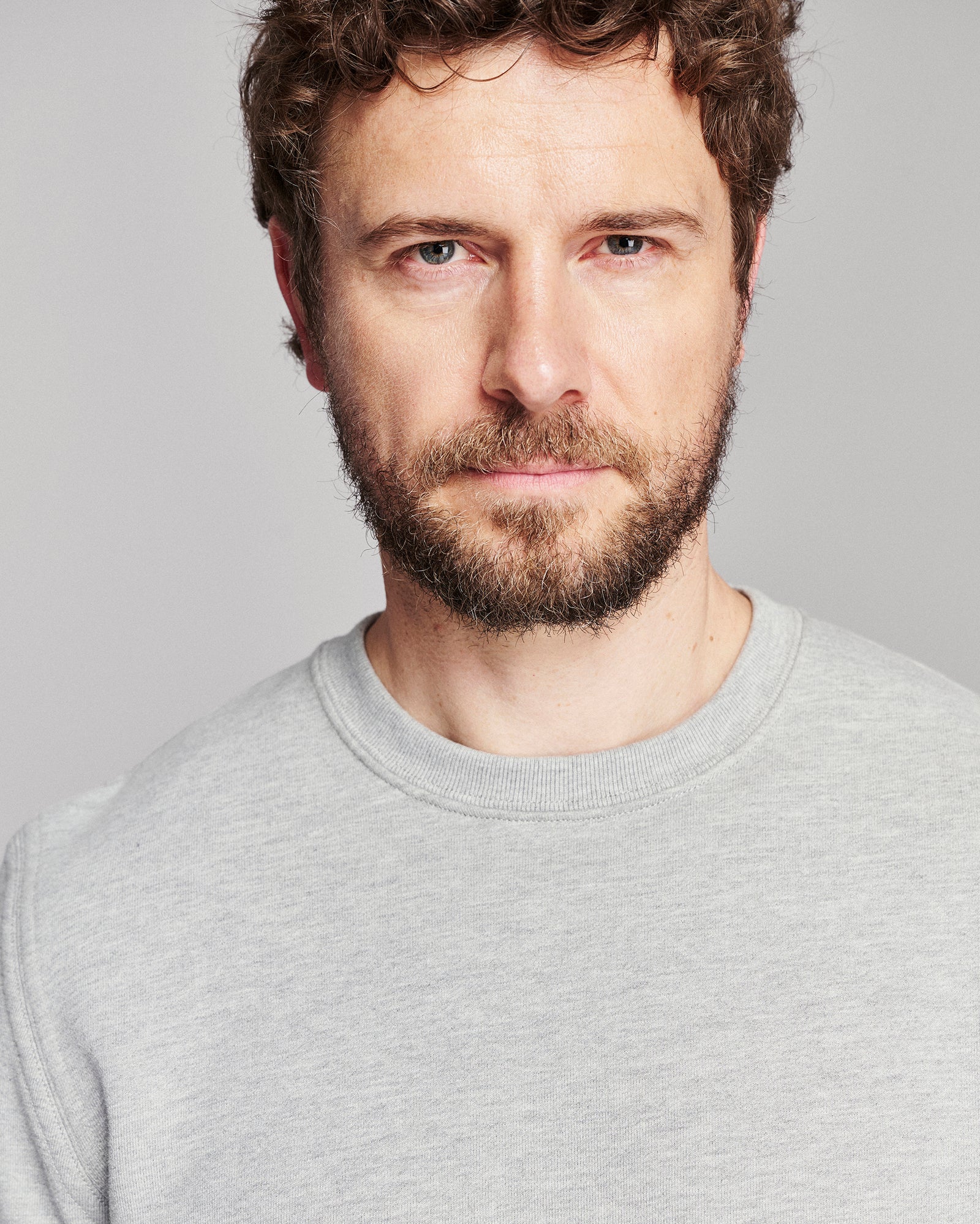 A close-up portrait of a man with curly brown hair and a beard, wearing a light gray crewneck sweatshirt against a solid gray background.
