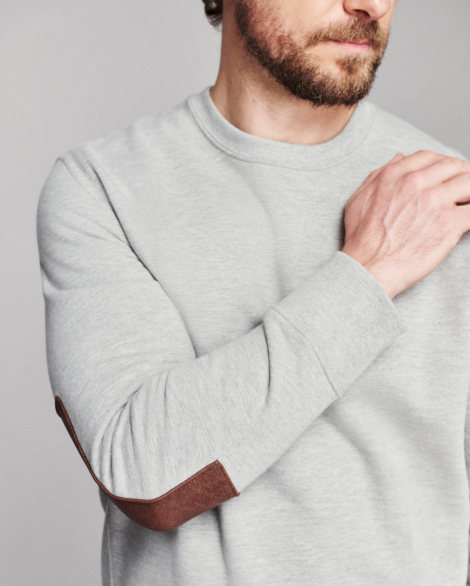 A close-up of a man wearing a light gray crewneck sweatshirt with brown elbow patches, shown from the chest up against a plain gray background.