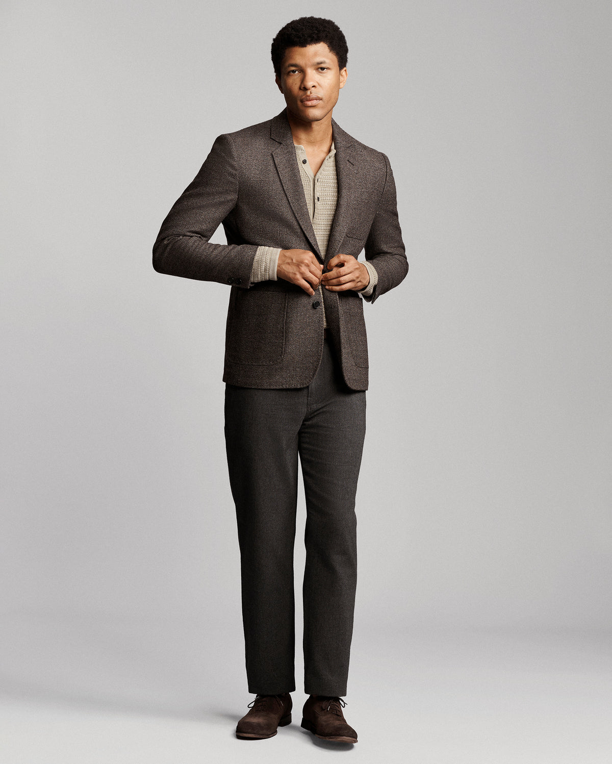 A man modeling a dark gray two-piece suit with a textured blazer and matching trousers, styled over a beige knit shirt and brown dress shoes, against a light gray studio background.