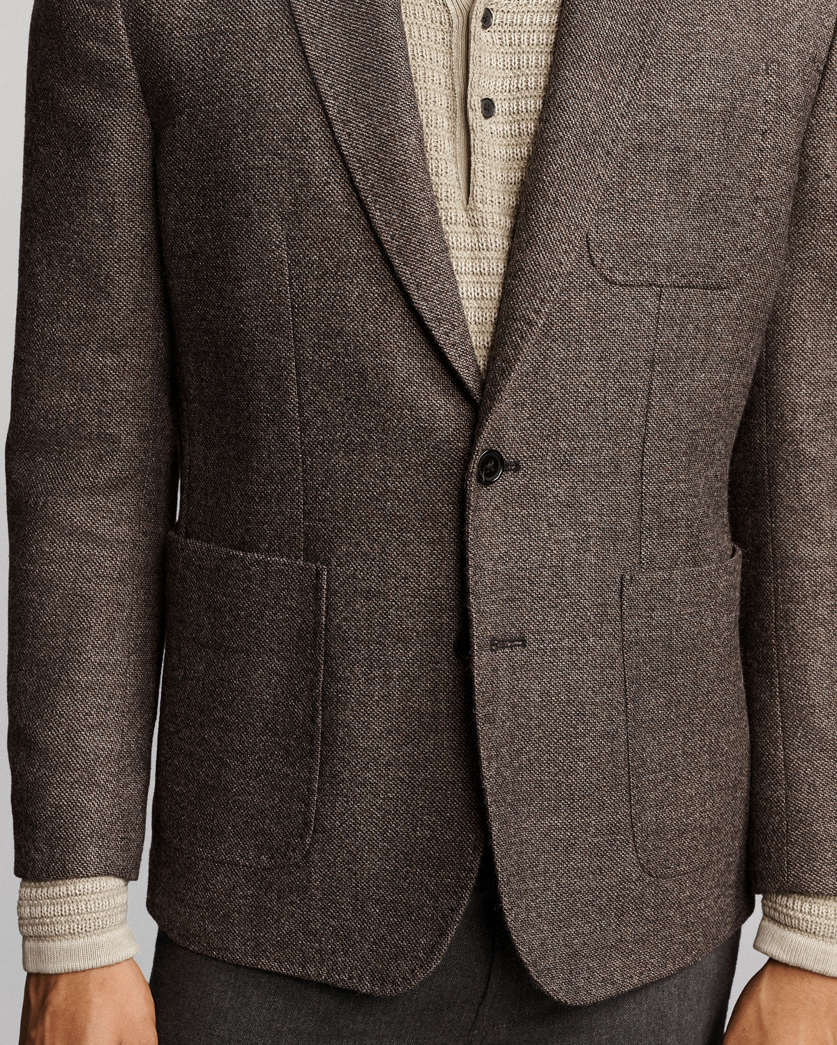 A close-up of a men's brown tweed blazer with notch lapels, a two-button closure, and front patch pockets, styled over a cream knit sweater and gray trousers.