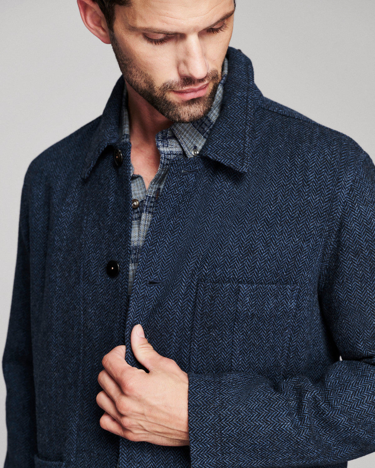 A close-up of a man wearing a dark navy blue herringbone wool coat over a plaid shirt, photographed against a neutral light gray background.