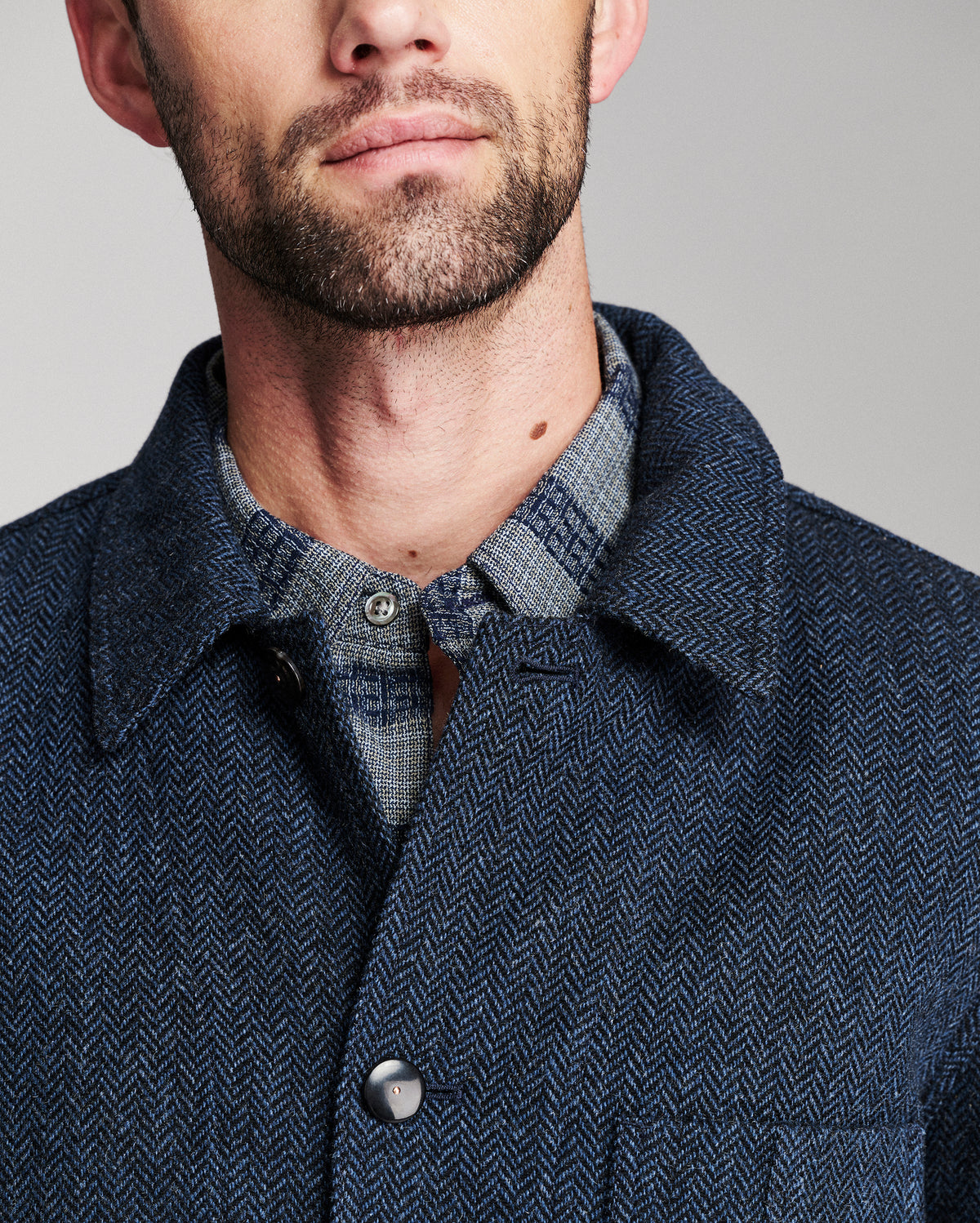Close-up of a man wearing a dark navy textured overshirt over a blue and gray plaid button-down shirt, against a neutral gray background.