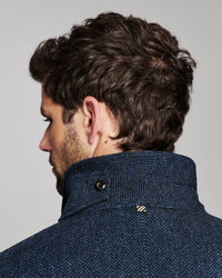 Close-up side profile of a man wearing a textured navy blue herringbone jacket with a high buttoned collar, against a light gray studio background.