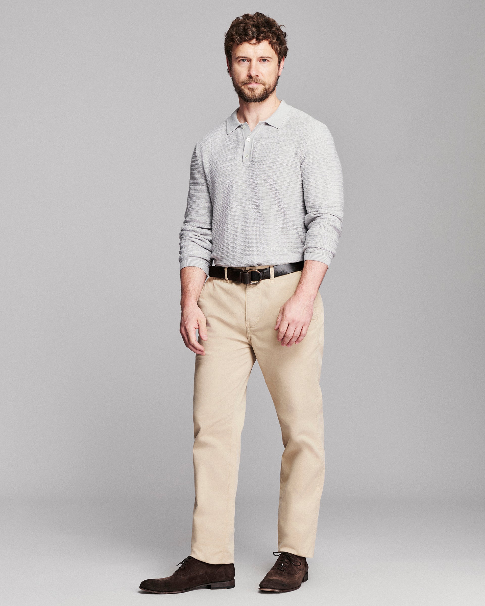 A man modeling a light gray long-sleeve polo shirt, beige trousers, a black belt, and dark brown dress shoes against a solid gray studio background.