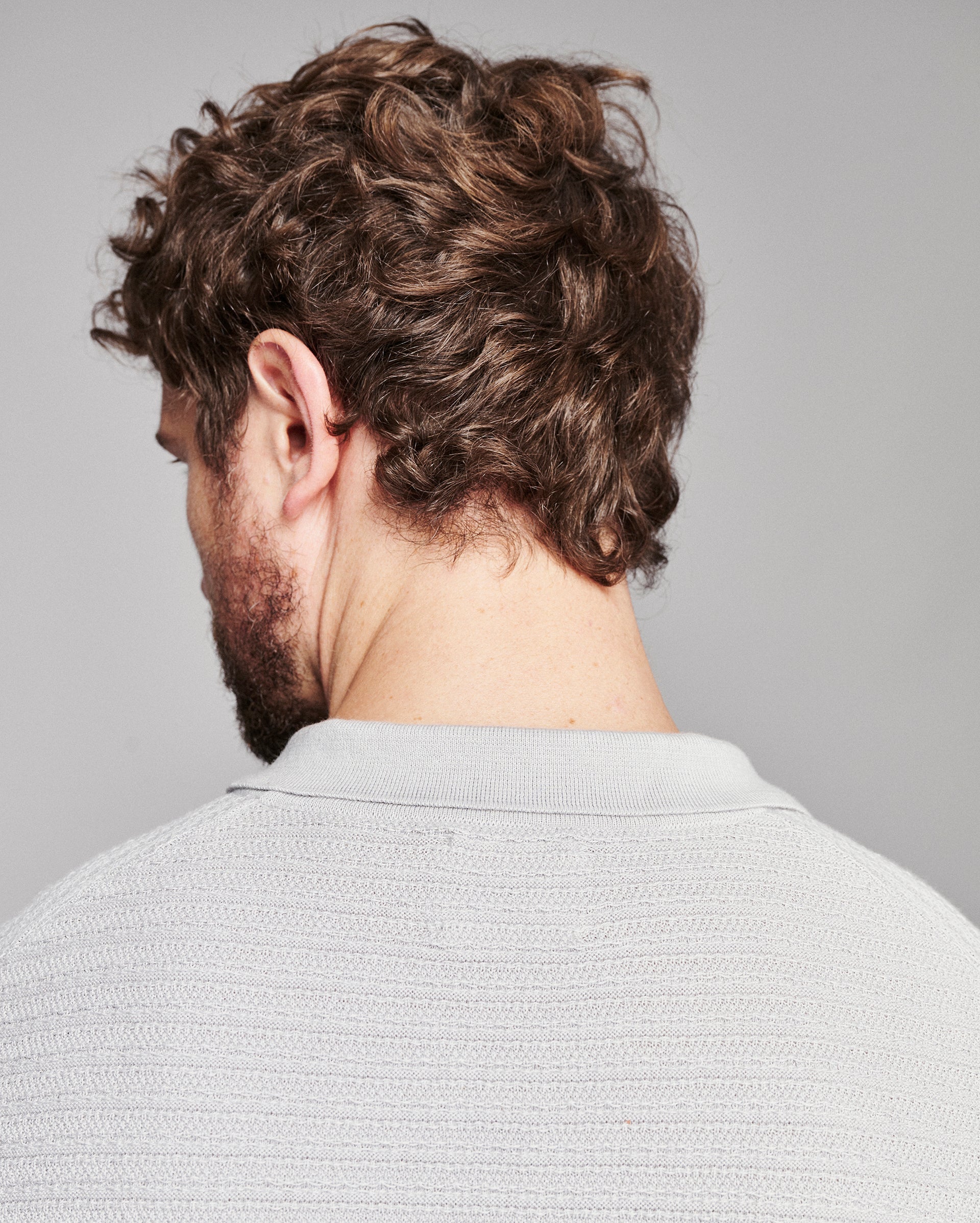 Rear view of a man with curly brown hair and a beard, wearing a light gray textured knit polo shirt against a solid gray background.