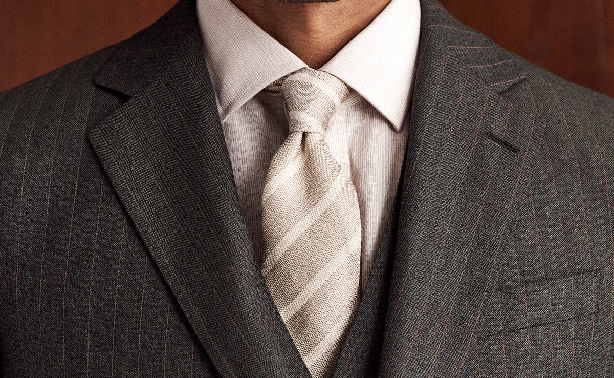 A close-up of a man wearing a dark gray pinstripe suit jacket, a light beige dress shirt, and a matching beige and white striped tie.