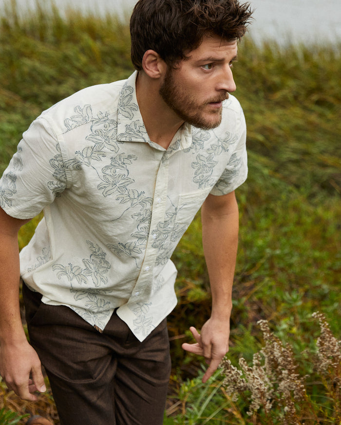 A man with short brown hair and a beard, wearing a light-colored, short-sleeve, floral button-up shirt and dark pants, stands outdoors surrounded by tall green grass.