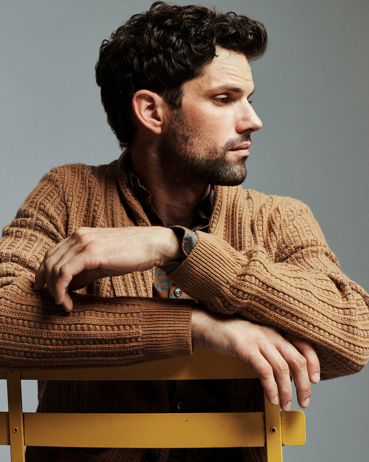 A man with dark curly hair and a beard wearing a brown cable-knit sweater, leaning on a yellow chair against a gray studio background.