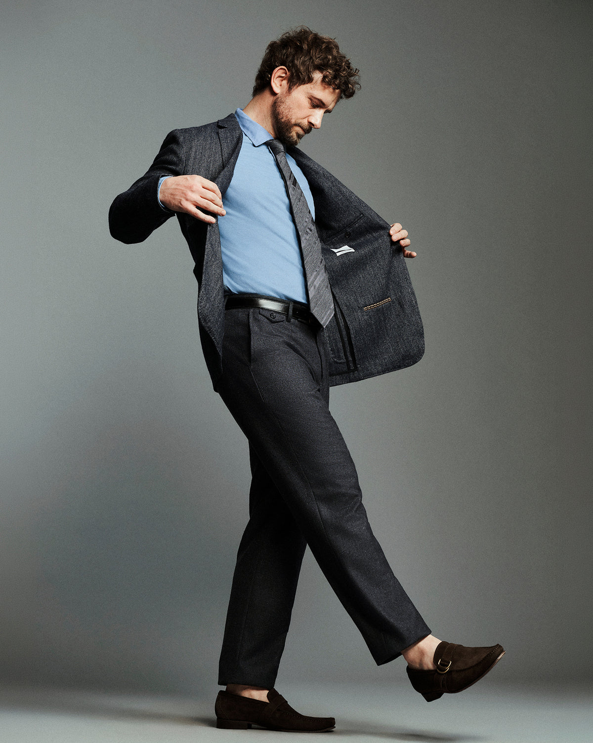 A man modeling a dark gray textured suit, featuring a light blue dress shirt, a gray patterned tie, and dark brown loafers, posing against a solid gray studio background.