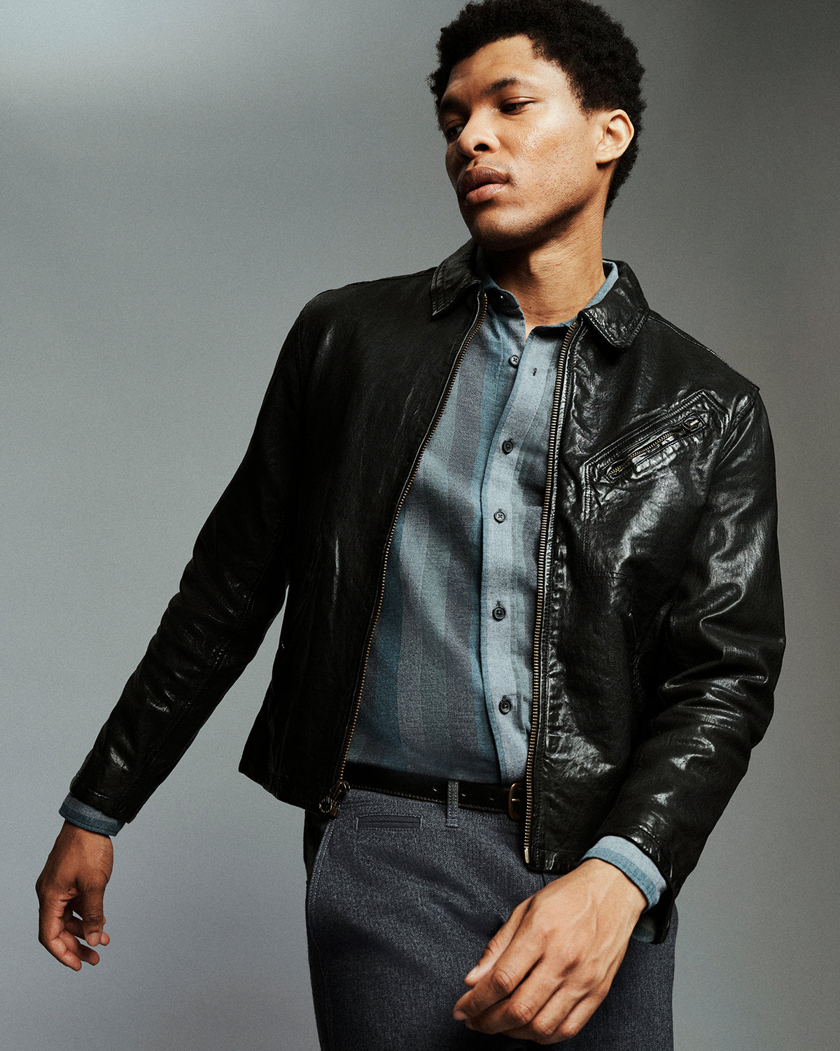 A man wearing a black leather jacket over a blue button-down shirt and gray trousers, photographed against a plain gray studio background.