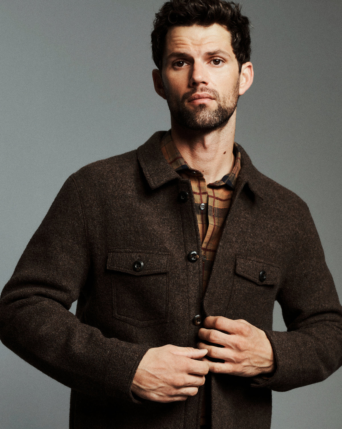 A man wearing a dark brown textured button-up overshirt over a brown and tan plaid shirt, posed against a solid gray studio background.