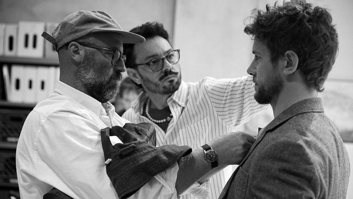 Black and white photograph of a man adjusting the collar of a suit jacket on another man, with a third man in the background wearing glasses and a striped shirt.