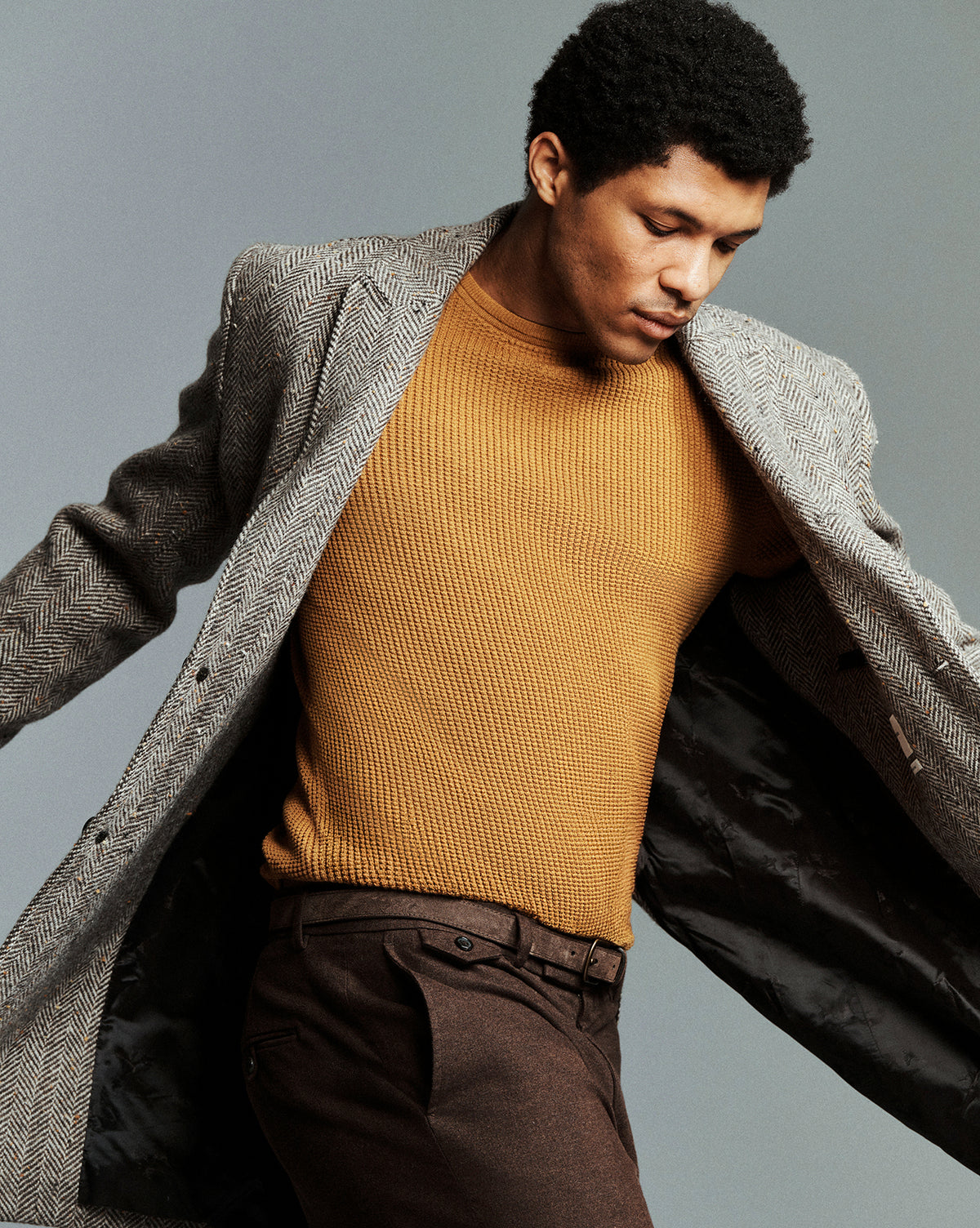 A man wearing a gray herringbone blazer over a mustard yellow ribbed knit sweater and brown trousers, posed against a solid gray studio background.