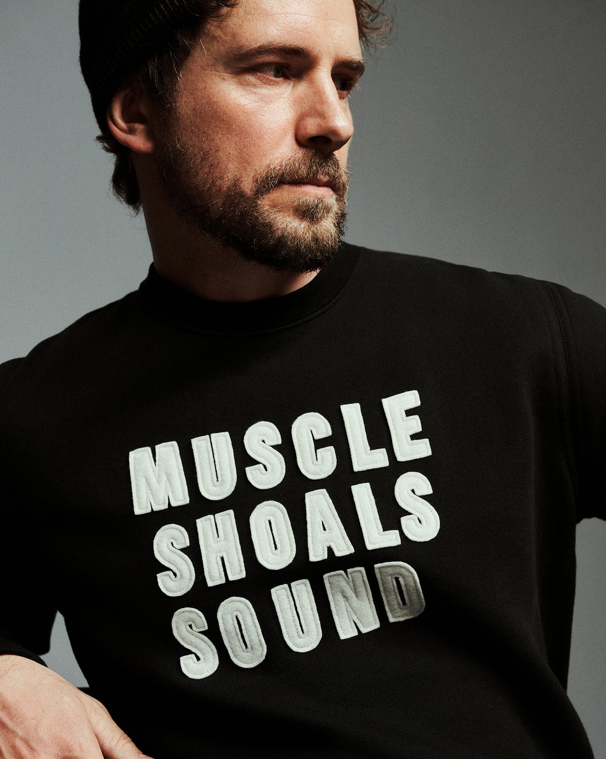 A man wearing a black crewneck sweatshirt with the text 'MUSCLE SHOALS SOUND' printed in large white and gray letters on the front, set against a dark gray studio background.