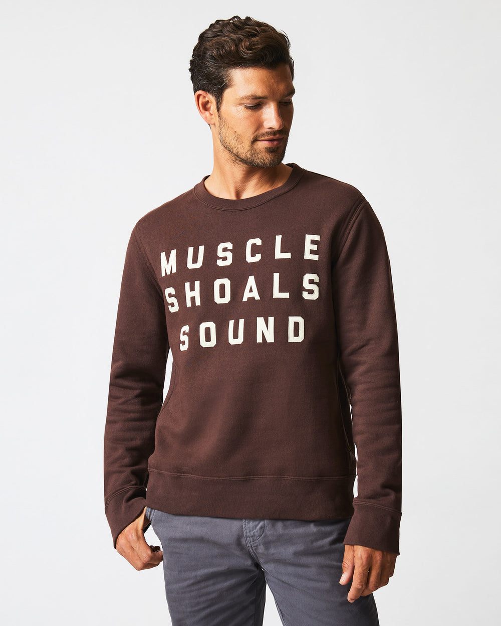 A man with short dark hair and a trimmed beard wears a brown sweatshirt with the white text MUSCLE SHOALS SOUND and gray pants, standing against a plain white background. He is looking down and to his left.