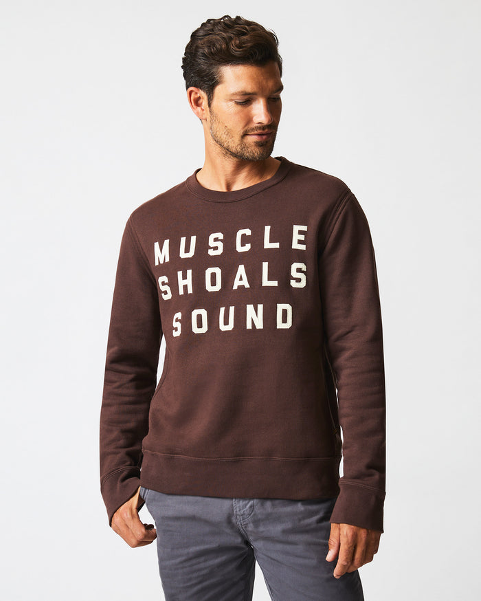 A man with short dark hair and a trimmed beard wears a brown sweatshirt with the white text MUSCLE SHOALS SOUND and gray pants, standing against a plain white background. He is looking down and to his left.