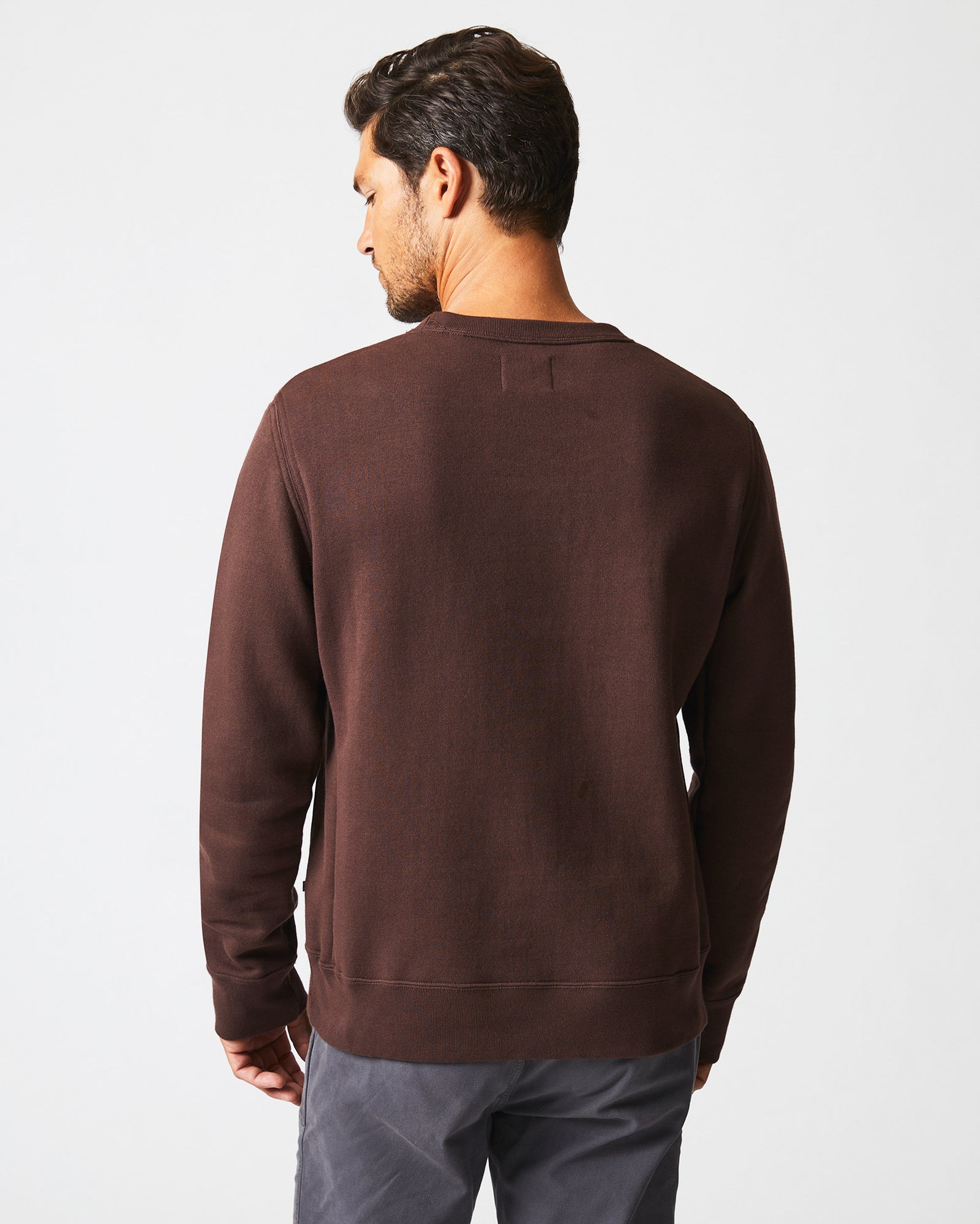 A man with short dark hair is standing and facing away from the camera, wearing a plain brown long-sleeve sweatshirt and gray pants against a white background.