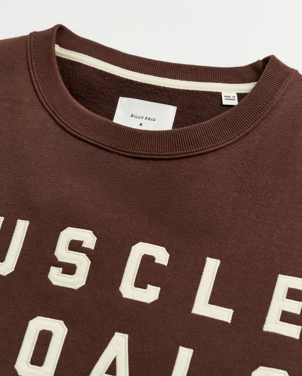 Close-up of a brown sweatshirt with beige, raised block letters spelling “MUSCLE GOALS” and a visible white tag at the neckline displaying the brand name “BILLY REID” and size “M.”.