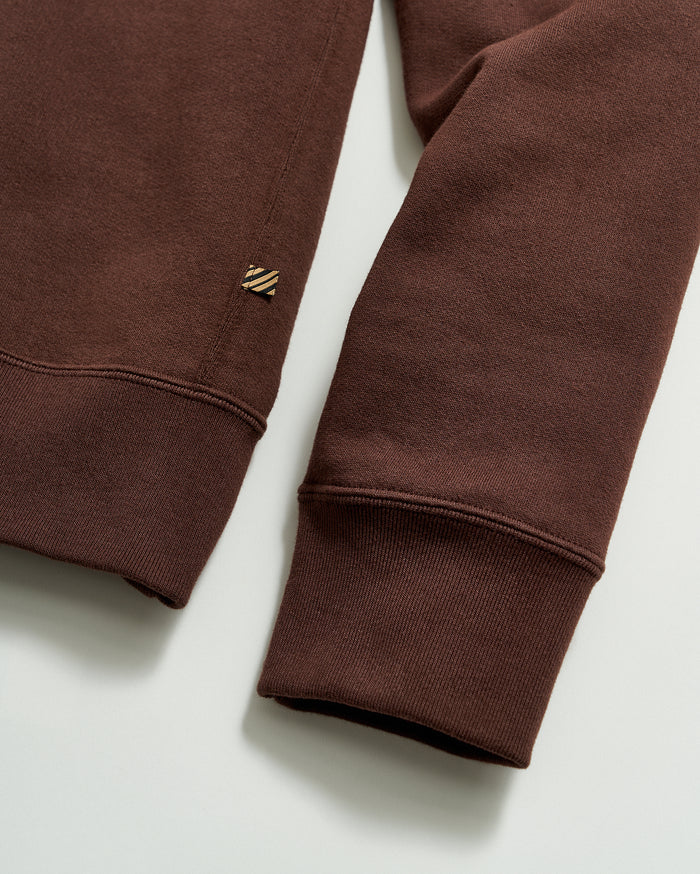 A close-up of a brown long-sleeve sweatshirt, showing the ribbed cuff and hem, with a small stitched logo tag near the hem. The fabric appears soft and comfortable.
