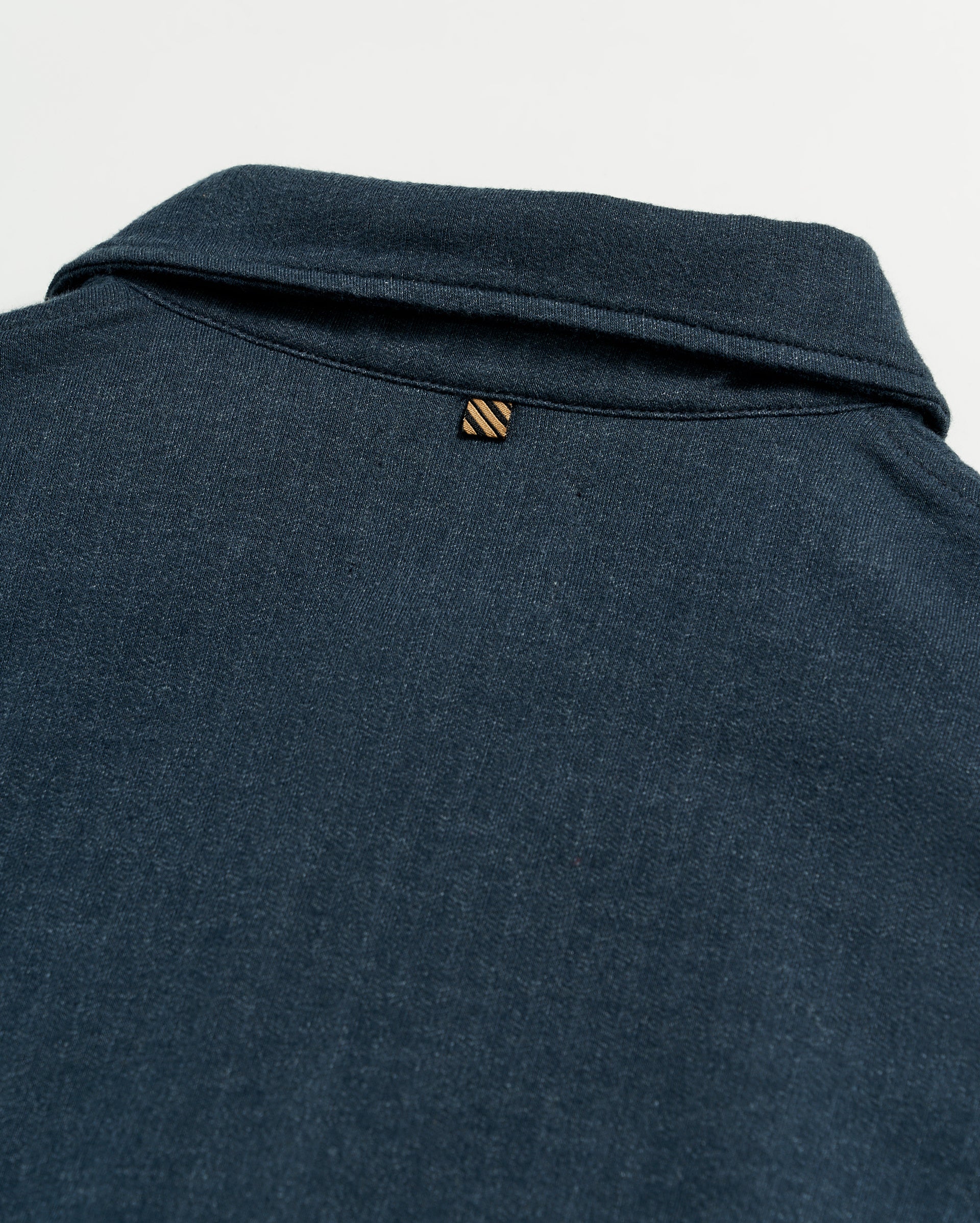 Close-up of the back of a dark blue collared shirt, showing a small, striped loop detail just below the collar on a plain white background.