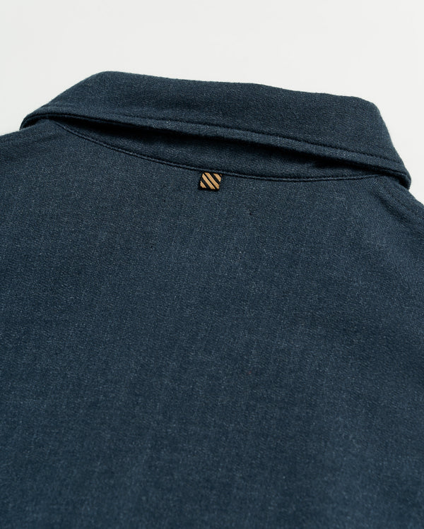 Close-up of the back of a dark blue collared shirt, showing a small, striped loop detail just below the collar on a plain white background.