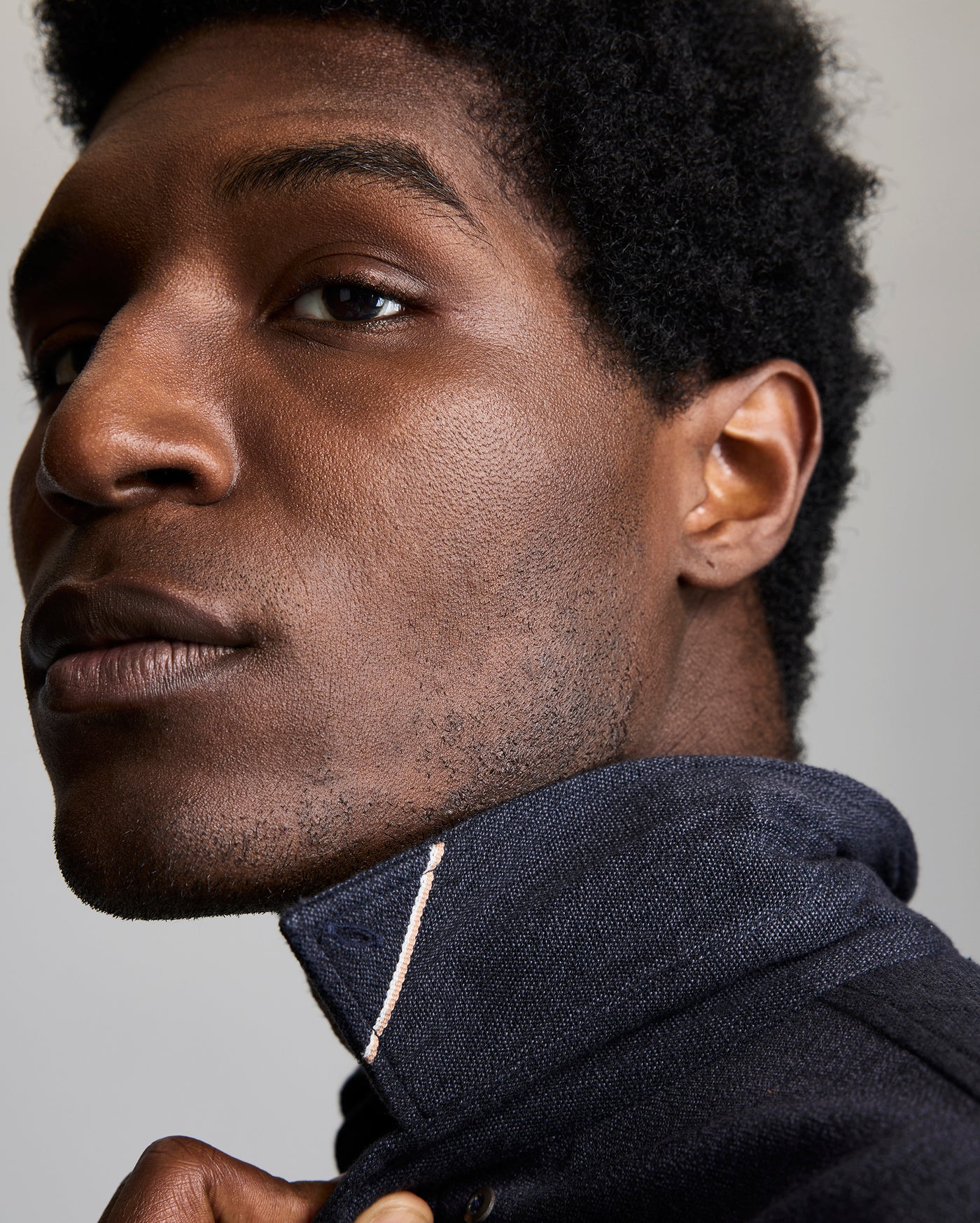 A close-up of a person with short curly hair wearing a navy blue collared jacket pulled up to their chin, looking slightly to the side with a neutral expression against a plain background.