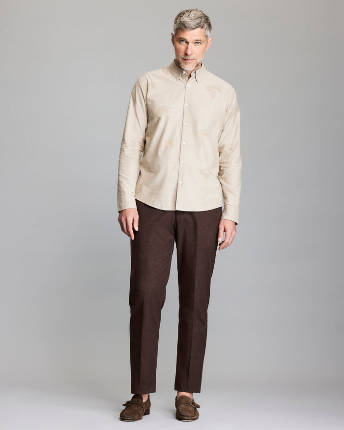 A man with gray hair wears a beige button-up shirt, dark brown pants, and brown loafers, standing against a plain gray background.