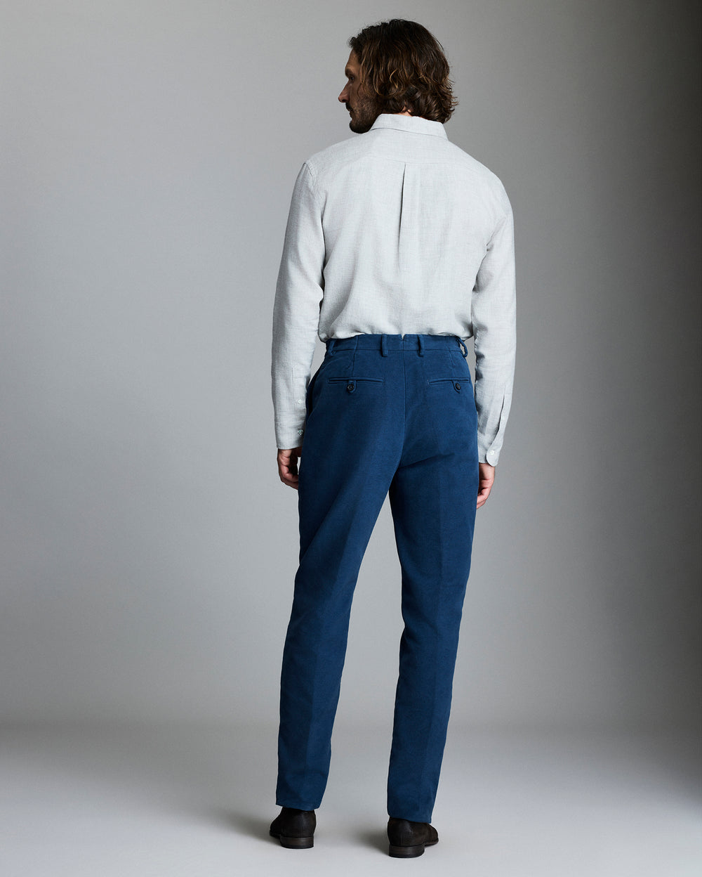 A man with shoulder-length hair stands facing away, wearing a light grey long-sleeve shirt tucked into blue trousers and black shoes, set against a plain grey background.