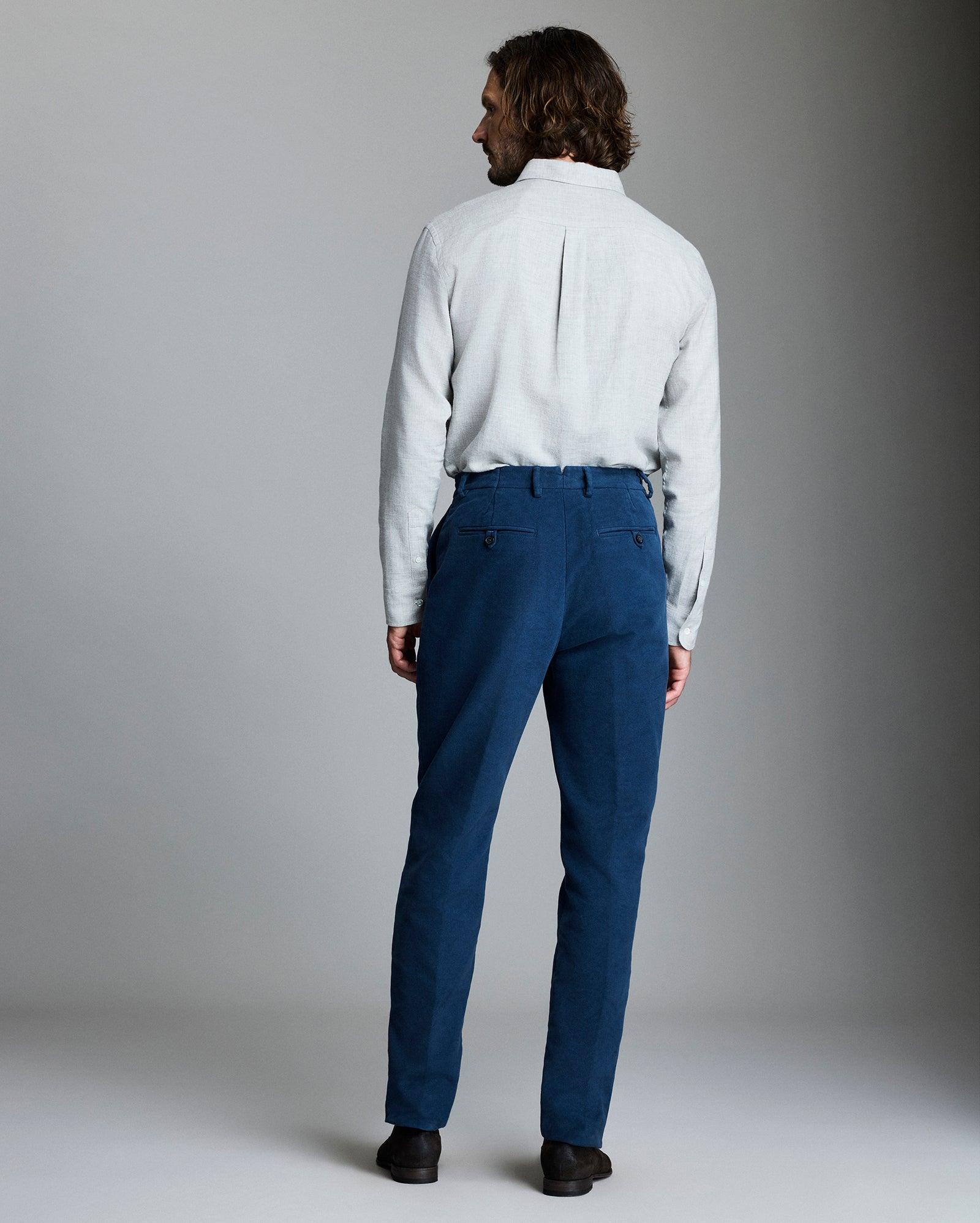 A man with shoulder-length hair stands facing away, wearing a light grey long-sleeve shirt tucked into blue trousers and black shoes, set against a plain grey background.