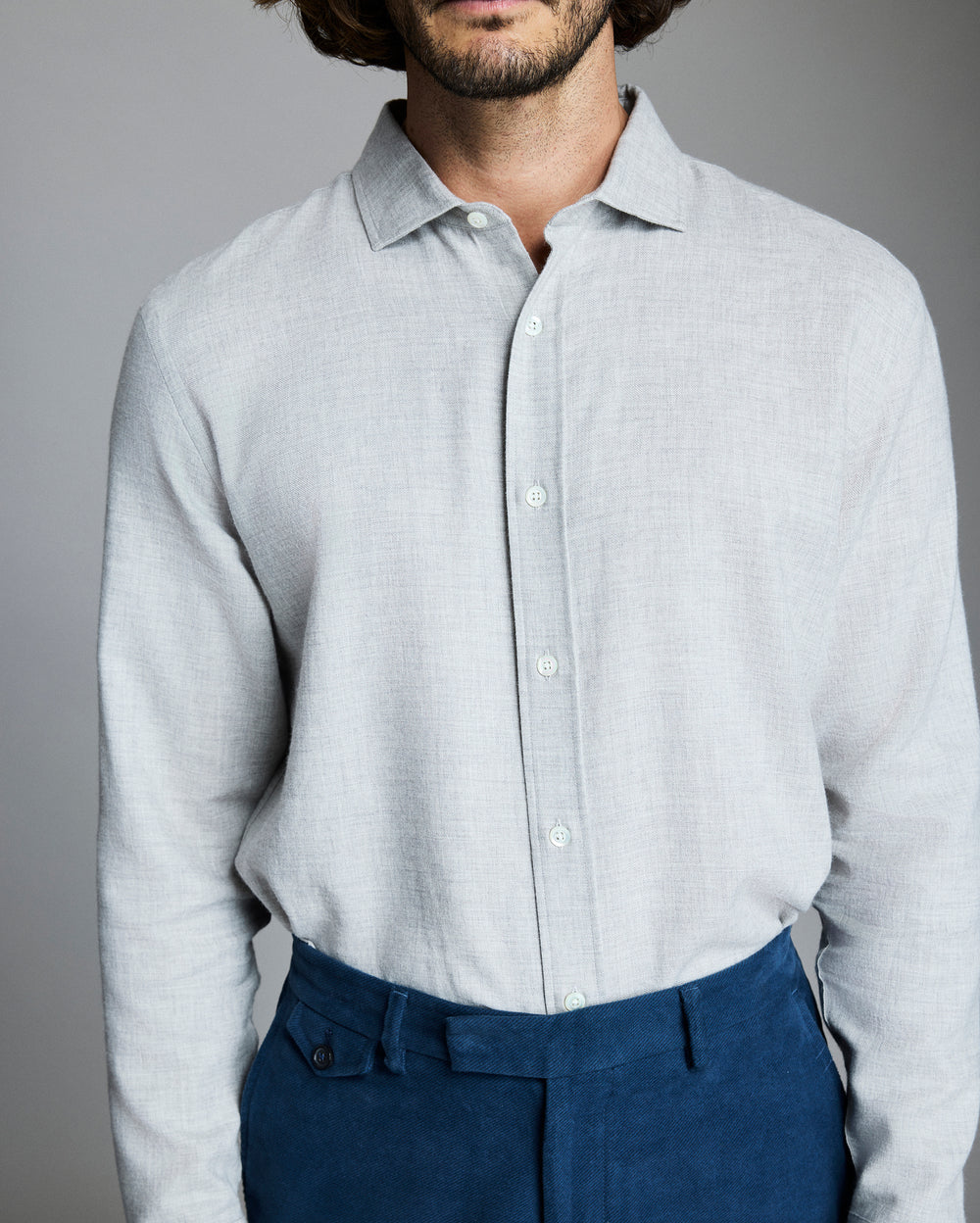 A man wearing a light gray button-up shirt and blue trousers stands against a plain gray background. His head is partially out of the frame, and his hands are at his sides.