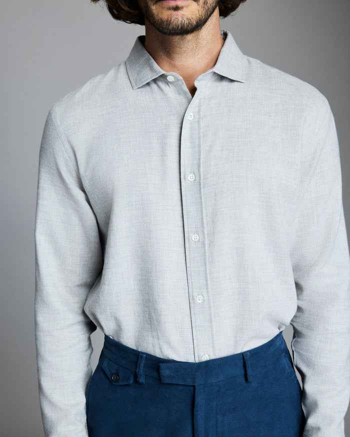 A man wearing a light gray button-up shirt and blue trousers stands against a plain gray background. His head is partially out of the frame, and his hands are at his sides.