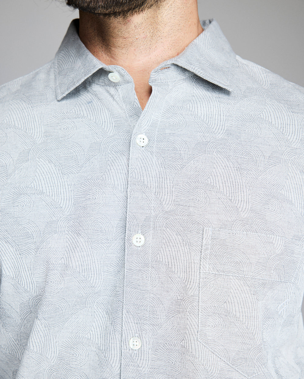 A close-up of a person wearing a light gray patterned button-up shirt with a collar and a chest pocket. The persons face is partially out of frame, showing only the lower part of their beard.