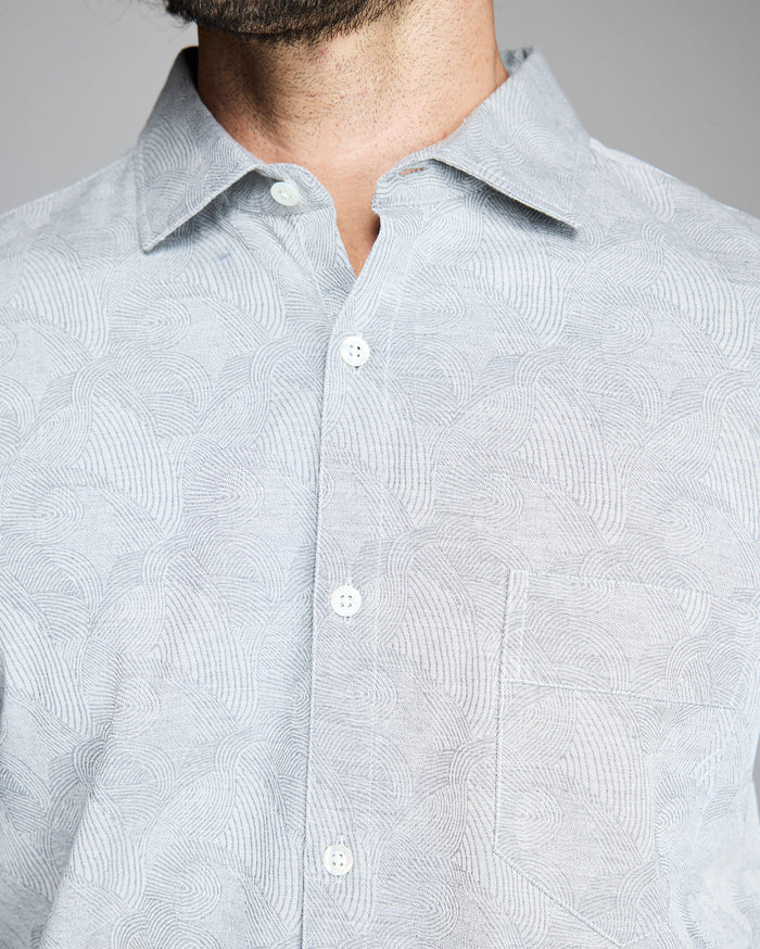 A close-up of a person wearing a light gray patterned button-up shirt with a collar and a chest pocket. The persons face is partially out of frame, showing only the lower part of their beard.