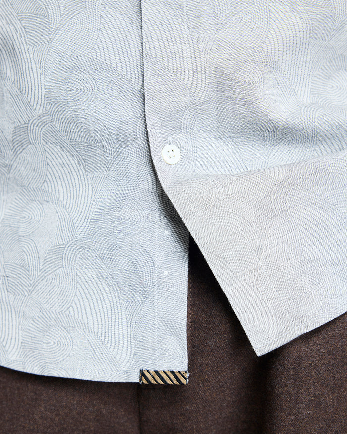 Close-up of a light gray patterned shirt with swirling lines, partially unbuttoned at the bottom, revealing dark brown pants underneath. A small striped fabric tag is visible near the shirt hem.