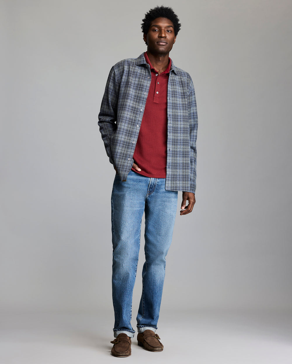 Man wearing a plaid shirt over a red polo shirt, blue jeans, and brown shoes on a gray background