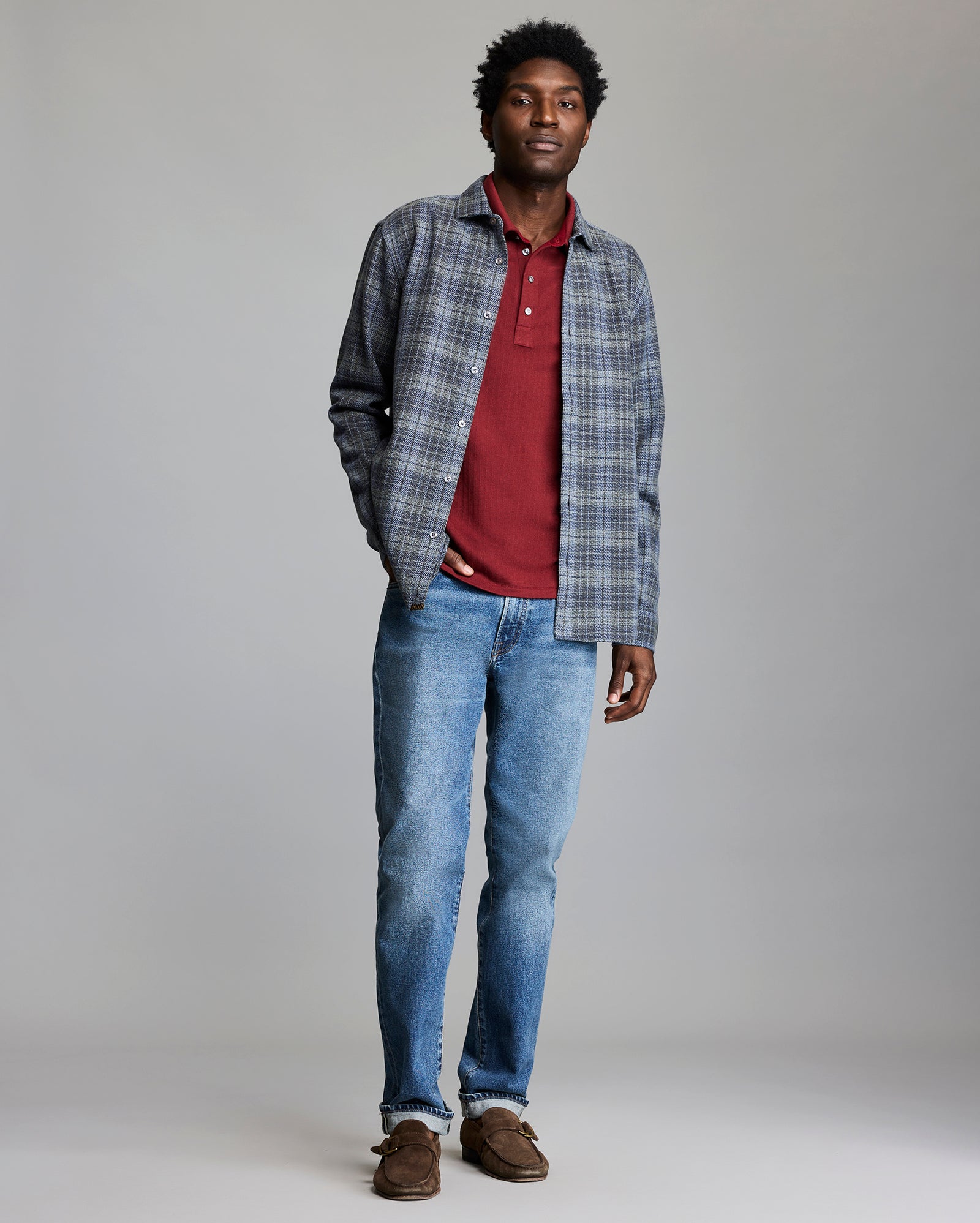 Man wearing a plaid shirt over a red polo shirt, blue jeans, and brown shoes on a gray background