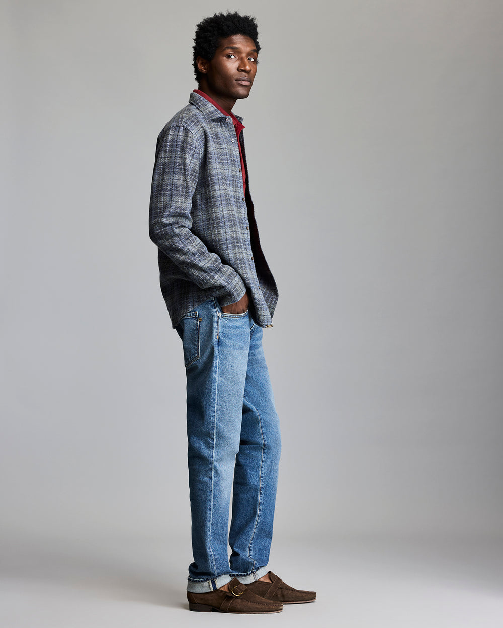 Man wearing a plaid shirt, red sweater, blue jeans, and brown shoes on a gray background
