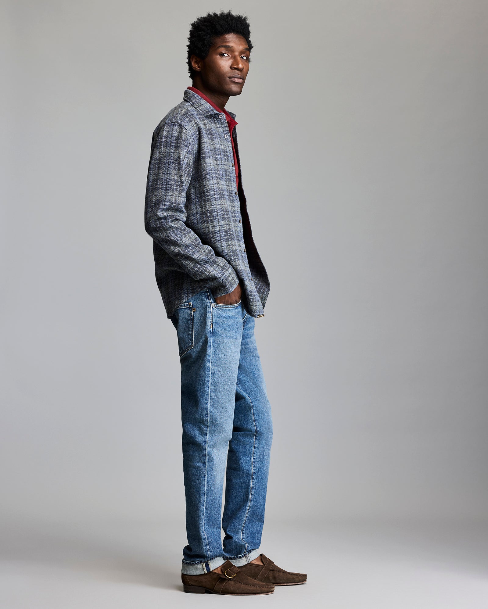 Man wearing a plaid shirt, red sweater, blue jeans, and brown shoes on a gray background
