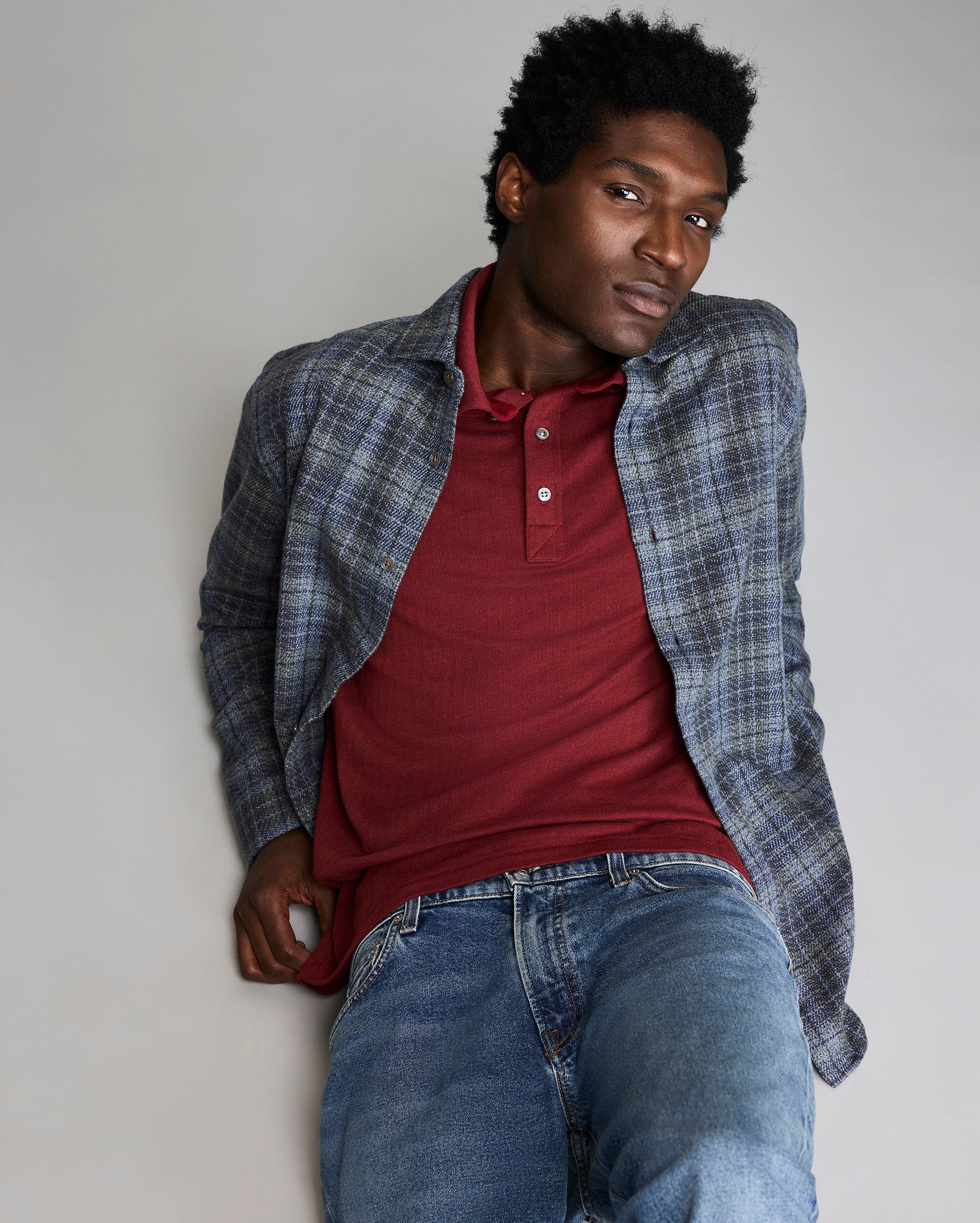 Man wearing a plaid shirt over a red polo shirt and blue jeans against a plain background