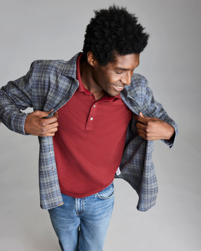 A man with short curly hair smiles while adjusting a gray plaid blazer over a red polo shirt and blue jeans, standing against a plain light background.