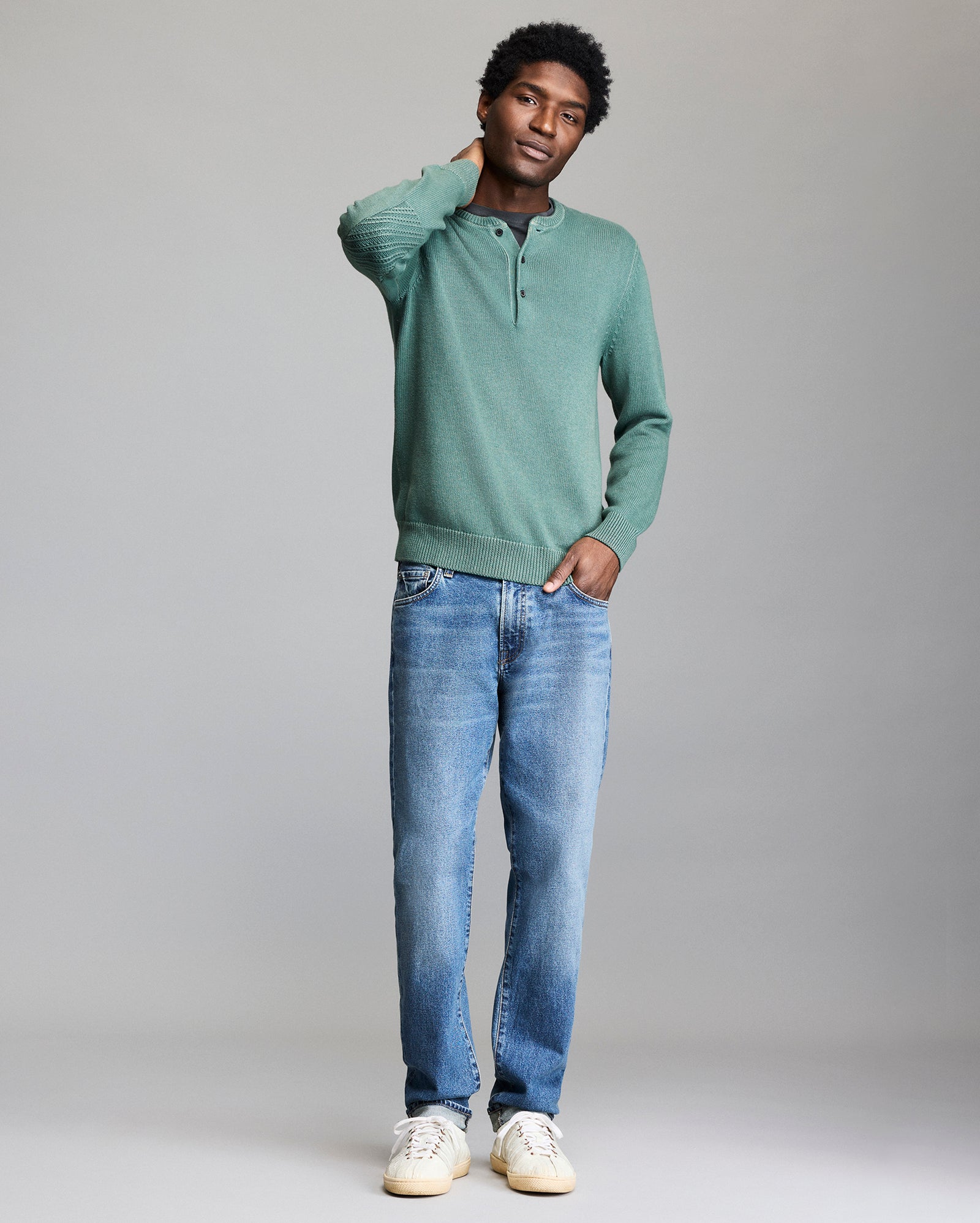 A person stands against a plain background, wearing a green long-sleeve henley shirt, blue jeans, and white sneakers, with one hand touching their neck and the other in a pocket.