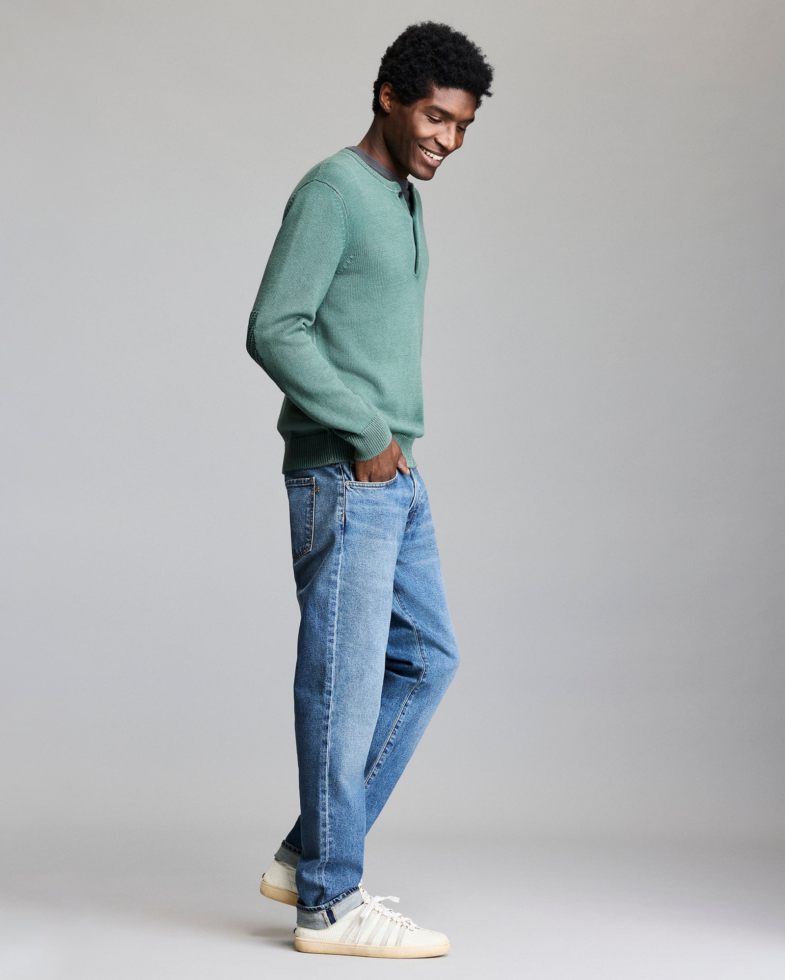 A man stands in profile smiling, wearing a green long-sleeve shirt, blue jeans, and white sneakers with stripes, against a plain gray background.